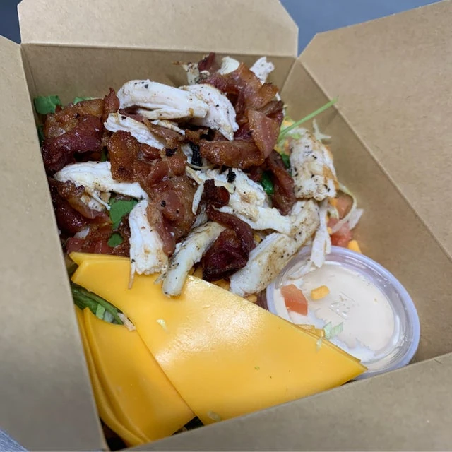 A mobile kitchen takeout box filled with a salad topped with grilled chicken, bacon, mango slices, and a side of white dressing.