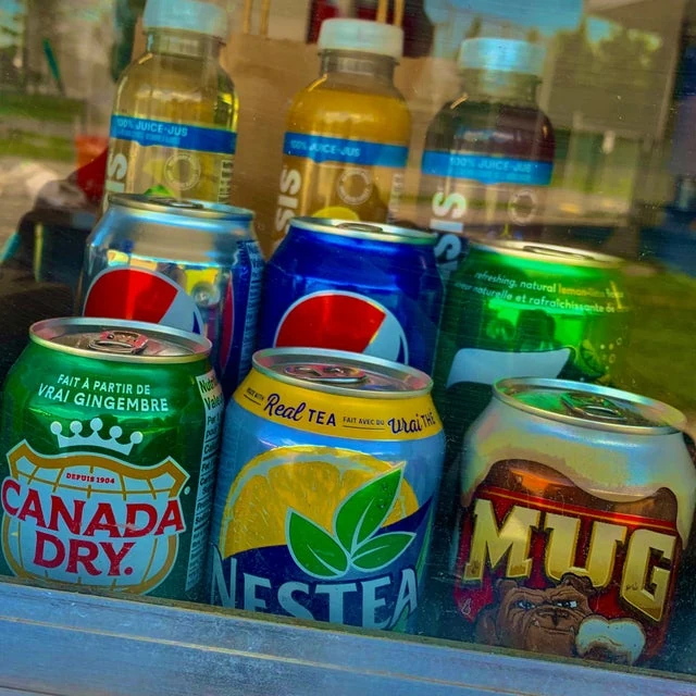 Various beverages including sodas and iced teas displayed behind a glass window of a mobile kitchen, reflecting greenery outside.