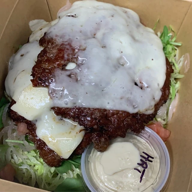 A fried chicken cutlet topped with melted cheese, served on a bed of lettuce in a mobile kitchen's cardboard takeout box with a side of sauce.
