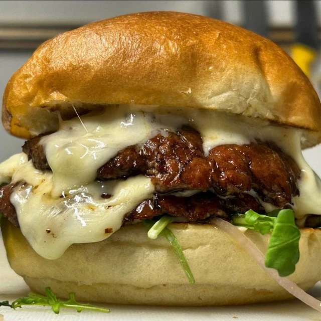Close-up of a burger with melted cheese, crispy chicken patty, lettuce, and red onions, prepared in a mobile kitchen and served in a shiny brioche bun.