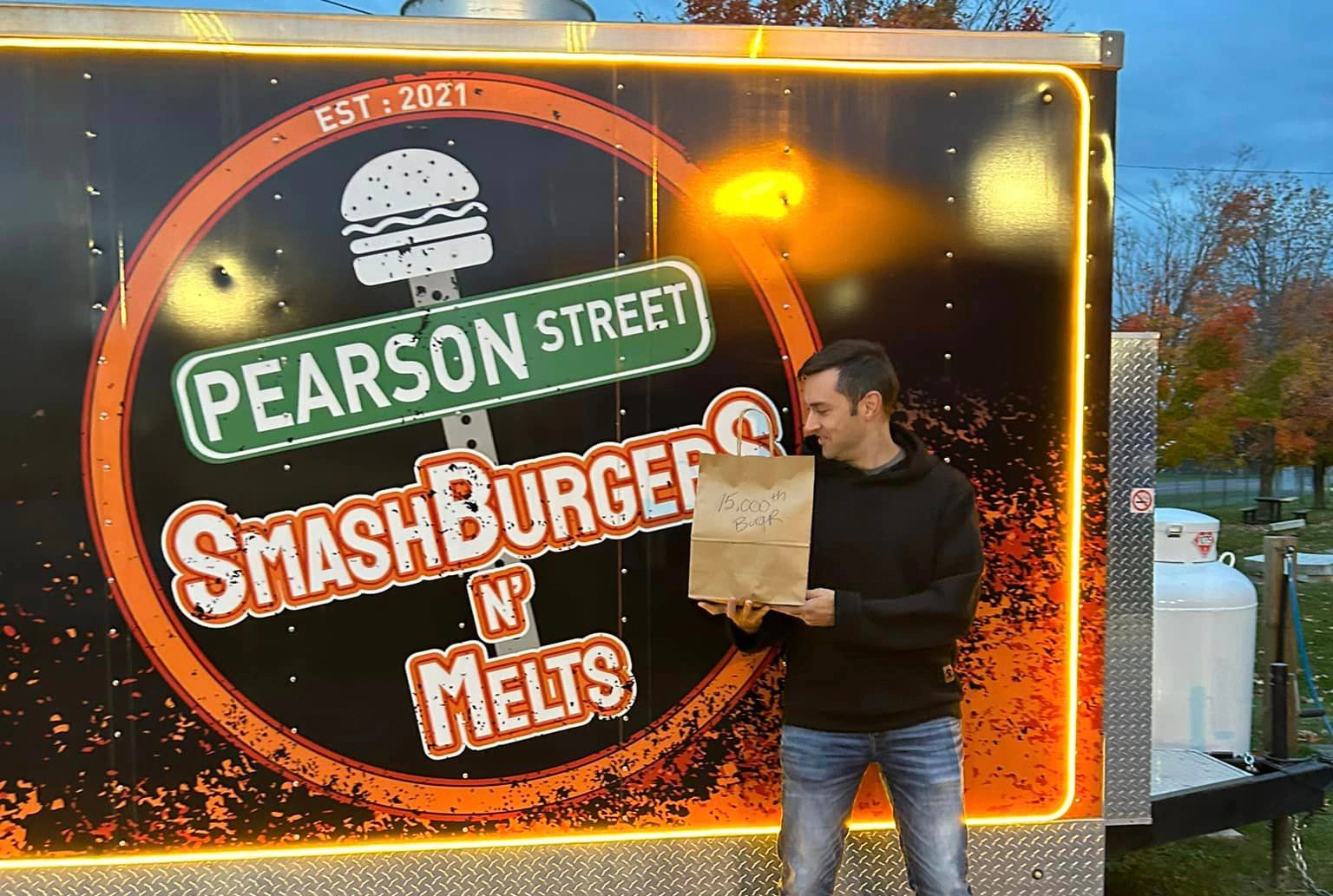 Man holding a paper bag stands in front of a "pearson street smashburger n melts" mobile kitchen during twilight, Contact