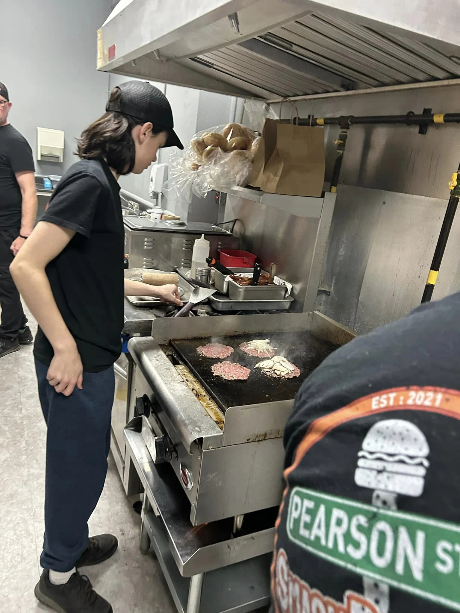 A kitchen worker grilling burgers on a flat top grill while another person observes in a commercial kitchen on Pearson Street.