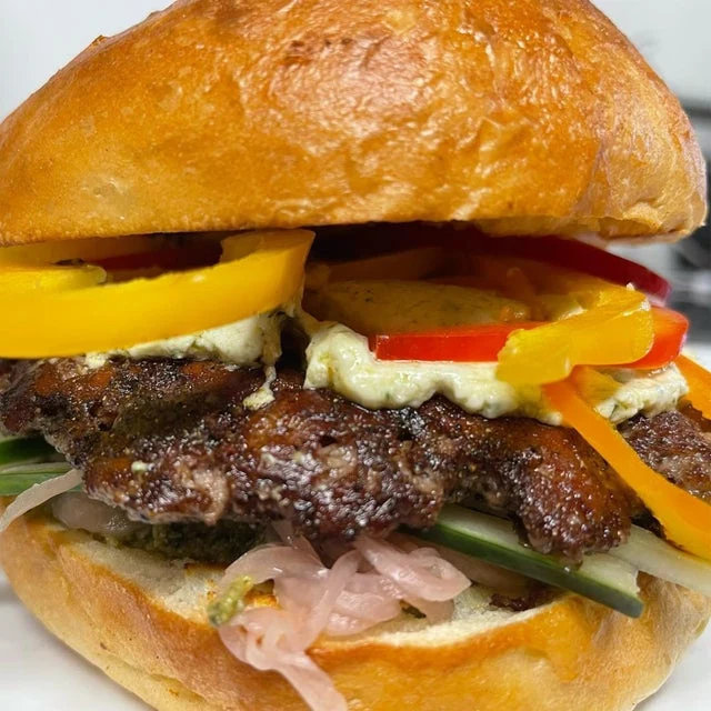 Close-up of a gourmet burger from a mobile kitchen, featuring a shiny bun, stacked with grilled meat, yellow and red bell peppers, onions, greens, and sauces.