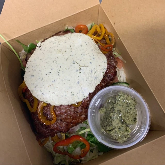 A takeout box from a mobile kitchen containing a burger topped with a large white sauce-covered patty, peppers, lettuce, and a side of green sauce.