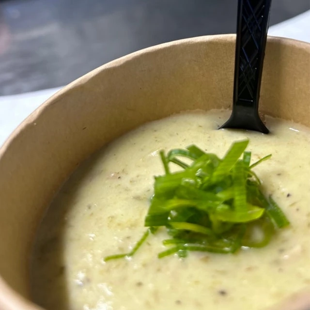 A bowl of creamy soup garnished with chopped green onions, prepared in a mobile kitchen and featuring a black spoon, presented on a white surface.