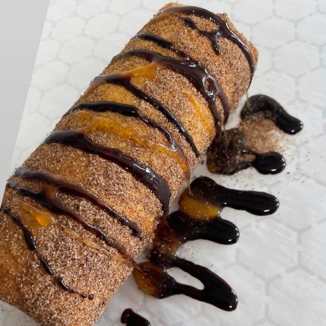 A cinnamon-coated pastry with a spiral cut, drizzled with caramel and chocolate sauce, served from a mobile kitchen on a white plate.