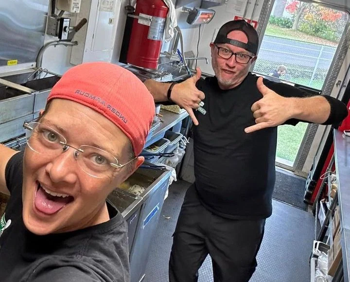 Two people smiling and posing with thumbs up inside a food truck kitchen on Pearson Street.